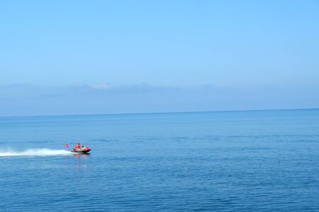 Two young men in lifejackets riding a white and yellow water scooter on the blue high seas, forming large waves, splashes and foam on the background cloudy sky.の写真素材