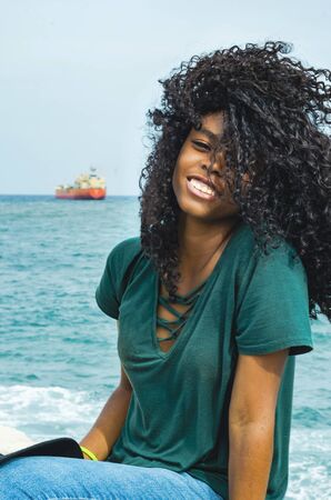 Young girl with black hair, laughing hair sitting on her back to the Caribbean Sea, enjoying a summer day with the wind in her hair, lifestyle portraitの写真素材