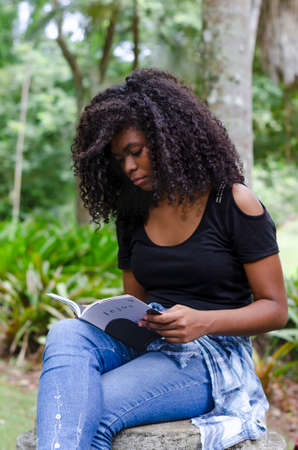 a young black woman between 20 and 30 years old sitting reading a book alone, in a park, surrounded by treesの写真素材