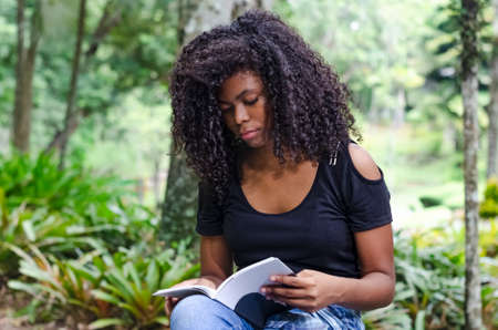 a young black woman between 20 and 30 years old sitting reading a book alone, in a park, surrounded by treesの写真素材