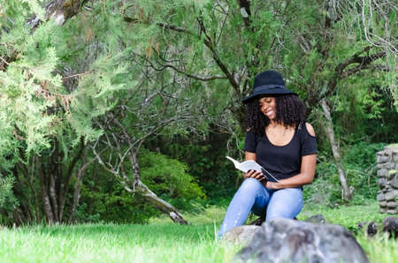 a young black woman between 20 and 30 years old sitting reading a book alone, in a park, surrounded...