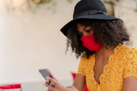 young black woman with curly hair, with red mask, yellow dress and black hat, checking her phoneの写真素材