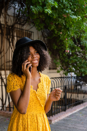 young black woman with curly hair, with red mask, yellow dress, using her smart phone happyの写真素材