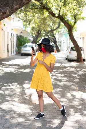 young black woman with curly hair, with red mask, yellow dress, using her smart phoneの写真素材