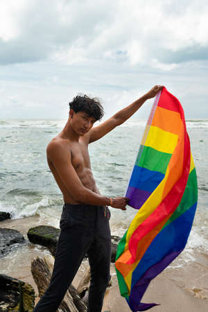 Portrait of young Hispanic gay boy looking at camera, holding LGBT flag - Focus on face vertical...