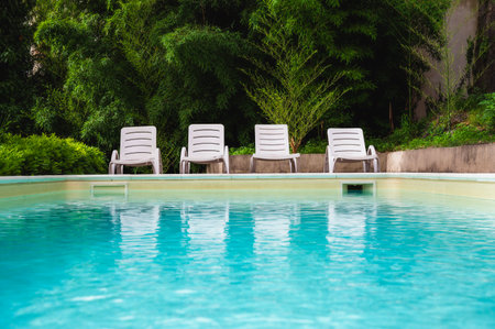 Swimming pool with stair and blue water. White Sunbeds in green lush garden by the blue swimming pool. Summer relax concept. Texture of water surface. Space for text.の写真素材