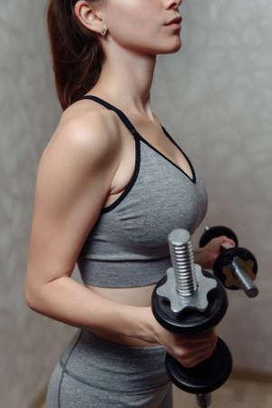 Close-up of a young woman in gray sportswear doing biceps exercise with dumbbells at homeの写真素材