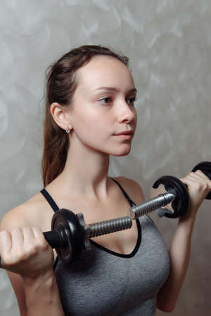 Close-up of a young woman in gray sportswear doing biceps exercise with dumbbells at home