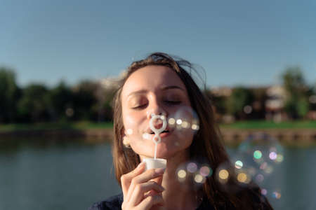 Gorgeous young brunette girl blowing soap bubbles in sunlit park.