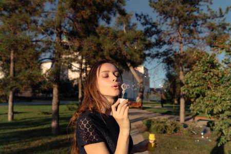 Gorgeous young brunette girl blowing soap bubbles in sunlit park.