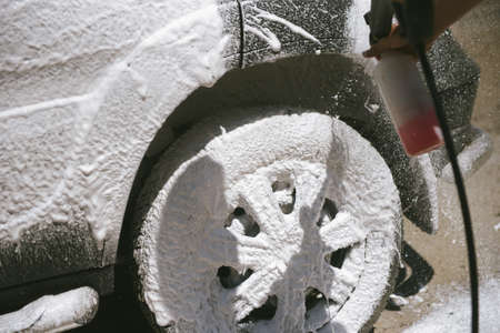 A man sprays a cleaning agent with a high pressure washer on a car. The car washes with a means of...