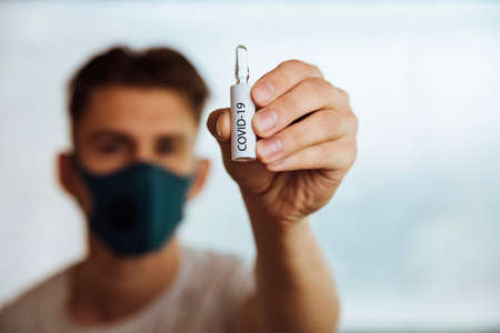 a man in a medical protective mask holds a test tube with the inscription covid-19の写真素材