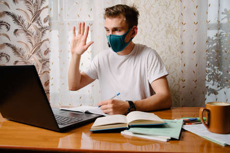 A young man in a face mask looks at the laptop screen and waves.A man works at home during quarantine on isolation.Freelance.の写真素材