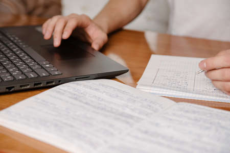 Student writing content on paper notebook with a pen while studying online course via laptop computer on wooden desk.Close upの写真素材