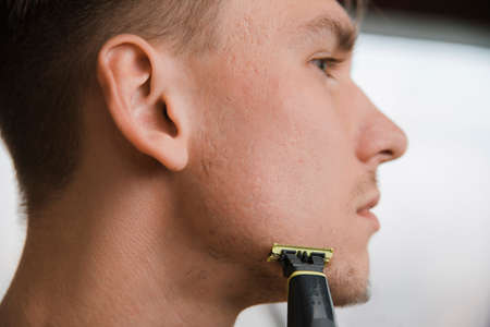 Close-up Portrait of handsome man shaving his beard with electric shaver in morning, against grey backgroundの写真素材