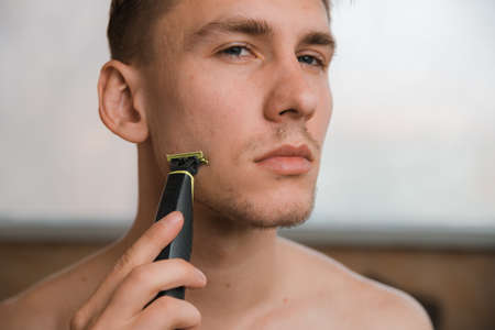Close-up Portrait of handsome man shaving his beard with electric shaver in morning, against grey...