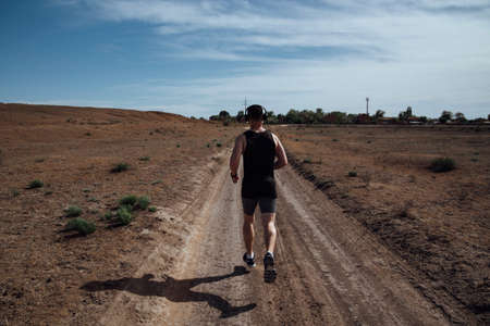 A man in sportswear and headphones runs along a dirt road in the summerの写真素材