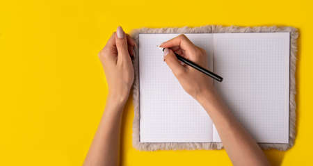 young woman hands hold opened notebook pages with black pen on yellow background.Top view, flat lay.Woman writes in a notebook,bannerの写真素材