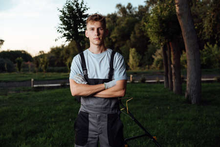 Young man in overalls posing with a lawn mower.Man mows the lawn with a lawn mower.Gardening and lawn mowing conceptの写真素材