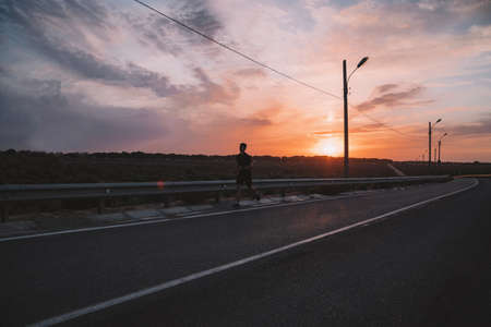 Attractive fit man running along modern bridge at sunset light, man doing workout outdoors, silhouette runner jogging overbridge road with amazing sunset on backgroundの写真素材