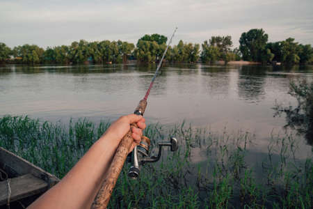 Fisherman with rod, spinning reel on the river bank. Sunrise. The concept of a rural getaway.Fisherman holds fishing rod close-up in the first personの写真素材
