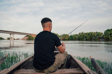 Young man fishing on a lake from the boat. Fisherman in the old, wooden rowboat and catching the fish on sunny day.の写真素材