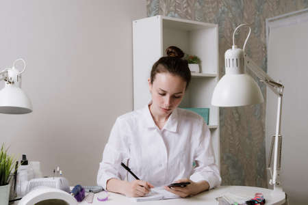 Beautiful girl manicurist in beauty salon.Manicure,pedicure and beauty concept.Manicurist sits at a workplace in a nail salon.Manicurist writes new clients to a notebookの写真素材