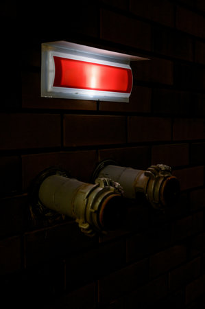 Dark interior with emergency lighting and utility pipes. Red emergency light above fire pipes on a brick wall.の写真素材