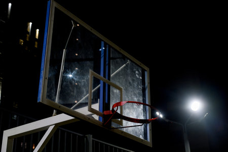 Close-up of a basketball backboard and red ring illuminated by a spotlight at night. Street basketball red hoop and backboard under bright artificial lighting.の写真素材