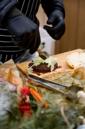 A chef in black gloves and an apron prepares a classic burger. Close-up of a juicy beef burger being prepared with patty and vegetables on a wooden board.の写真素材