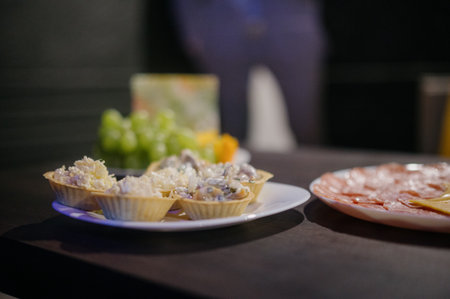 Several tartlets with salad on a plate and cold meats for a buffet close-up. Close-up of banquet appetizers with grapes in the background on a dark backgroundの写真素材