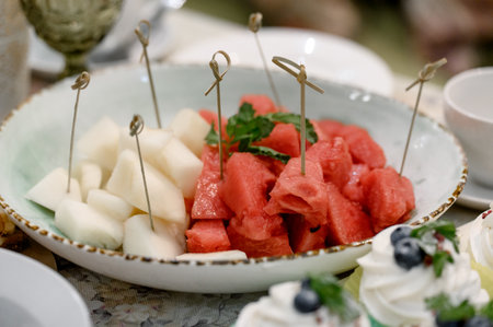 Fruit platter of watermelon and melon cubes with snack skewers. Fresh fruits for a buffet on a plate with berries and mint close-upの写真素材