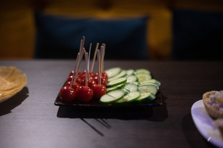 A close-up of a plate with assorted vegetables for canapes on skewers. Appetizer with cherry tomatoes and sliced cucumbers on skewers for a buffet on a dark tableの写真素材