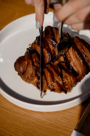 A woman's hands cut a chocolate croissant with a knife in a cafe. A close-up of a woman eating a fresh croissant dipped in dark chocolate.の写真素材