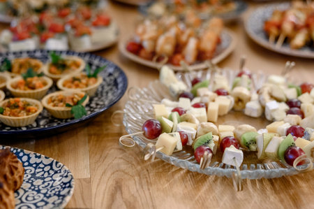 A platter of fruit and cheese skewers and tartlets on a wooden table during a banquet. Close-up of serving light appetizers on skewers with kiwi cheese and grapes on a clear plate.の写真素材
