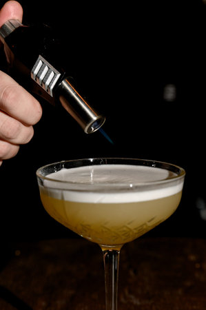 A bartender burns the surface of a cocktail foam with a gas torch in a dark bar. A bartender's hand...