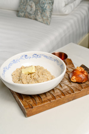 Oatmeal in a white ceramic bowl on a wooden stand with jam and a bun. Breakfast with oatmeal bun and red sauce on a carved wooden board in a bright interiorの写真素材