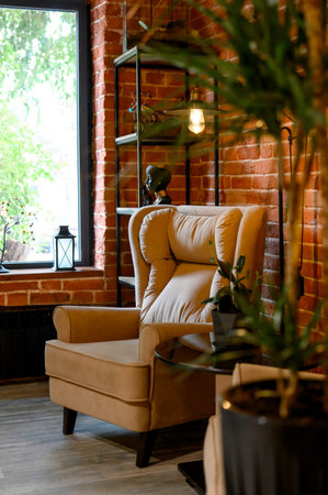 A beige soft armchair by the window against a brick wall in a modern interior.A cozy reading area with an armchair and black shelving in a brick roomの写真素材