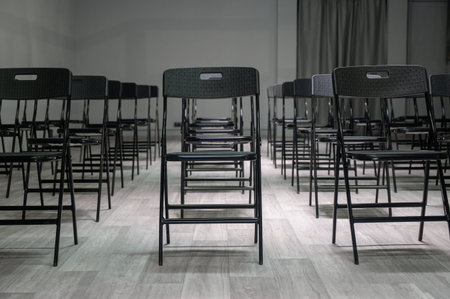 Rows of empty black folding chairs in a bright room. Empty black plastic chairs stand in rows on a...