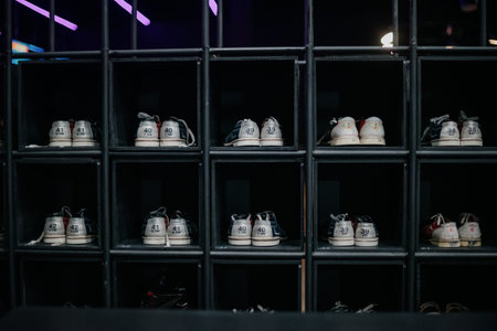 Rows of bowling shoes line the shelves at a sports equipment rental shop. A rack of pairs of special bowling shoes of different sizes in the compartments of a dark cabinetの写真素材