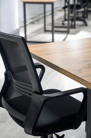 A black mesh office chair sits next to a wooden desk in a bright room. Close-up of the back of an ergonomic chair and the edge of the desk top in a modern office interiorの写真素材
