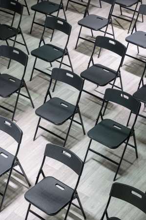 Rows of empty black folding chairs in a bright room. Empty black plastic chairs stand in rows on a light laminate floor in a classroom.の写真素材