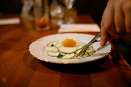 Close-up of a plate featuring cucumber slices and a garnish, with a hand holding a fork and knife,...