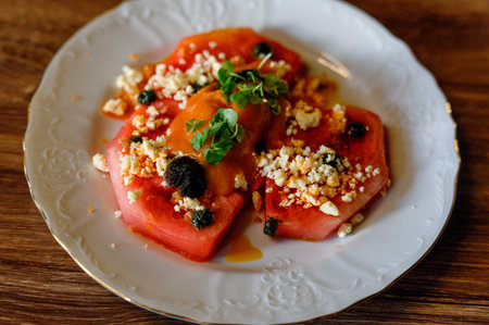 Fresh watermelon slices topped with crumbled feta cheese, herbs, and black olives arranged on a decorative white plateの写真素材