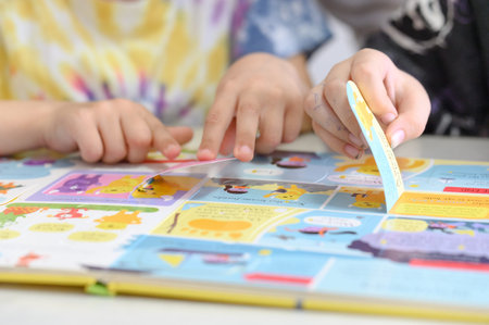 Two children engage with a vibrant storybook featuring illustrations and interactive flaps, placed on a light-colored table in a cozy settingの写真素材