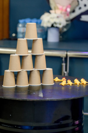 A pyramid of brown paper cups is arranged on a black table, with small rubber ducks placed in front,...