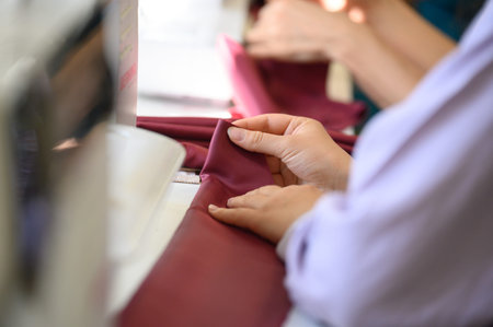 Close-up of hands folding maroon fabric near a sewing machine in a workshop, with another person's...