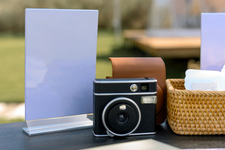 A camera sits next to a blank sign and a wicker basket on a table in an outdoor setting, with greenery visible in the backgroundの写真素材