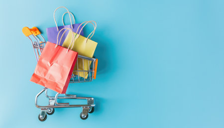A miniature shopping cart with colorful shopping bags on a light blue background.の素材
