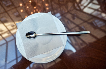 Illuminated glass restaurant roof reflecting in glassy table surface and polished stainless steel spoon on white paper towel on ceramic dishの写真素材